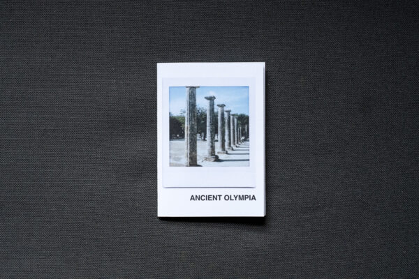 Photograph of an A7 zine on a dark grey fabric background. The zine has the word 'ANCIENT OLYMPIA' in the bottom right corner and an Instax Square print on the cover that shows columns in the Ancient Olympia archaeological site and blue sky above.