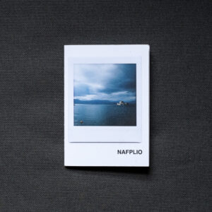 Photograph of an A7 zine on a dark grey fabric background. The zine has the word 'NAFPLIO' in the bottom right corner and an Instax Square print on the cover that shows the sea with a fort on the horizon, mountains in the background and stormy sky above.