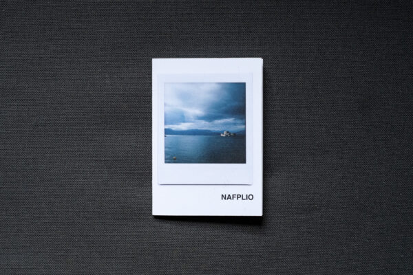 Photograph of an A7 zine on a dark grey fabric background. The zine has the word 'NAFPLIO' in the bottom right corner and an Instax Square print on the cover that shows the sea with a fort on the horizon, mountains in the background and stormy sky above.