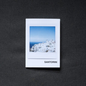 Photograph of an A7 zine on a dark grey fabric background. The zine has the word 'SANTORINI' in the bottom right corner and an Instax Square print on the cover that shows white buildings on the mountain top in Santorini and blue sky above.