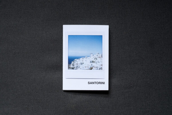 Photograph of an A7 zine on a dark grey fabric background. The zine has the word 'SANTORINI' in the bottom right corner and an Instax Square print on the cover that shows white buildings on the mountain top in Santorini and blue sky above.