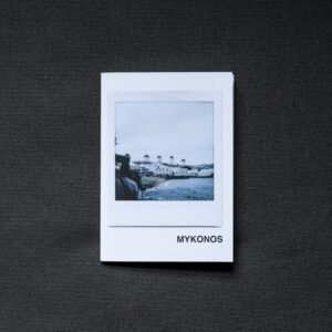 Photograph of an A7 zine on a dark grey fabric background. The zine has the word 'MYKONOS' in the bottom right corner and an Instax Square print on the cover that shows windmills on the horizon, sea in the foreground and cloudy sky above.