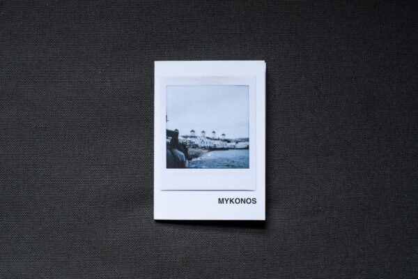 Photograph of an A7 zine on a dark grey fabric background. The zine has the word 'MYKONOS' in the bottom right corner and an Instax Square print on the cover that shows windmills on the horizon, sea in the foreground and cloudy sky above.