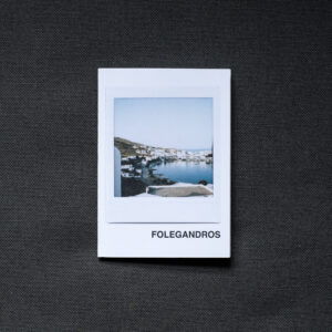 Photograph of an A7 zine on a dark grey fabric background. The zine has the word 'FOLEGANDROS' in the bottom right corner and an Instax Square print on the cover that shows the harbour in Folegandros with buildings around the sea and hazy sky above.