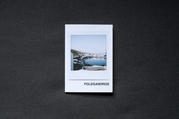 Photograph of an A7 zine on a dark grey fabric background. The zine has the word 'FOLEGANDROS' in the bottom right corner and an Instax Square print on the cover that shows the harbour in Folegandros with buildings around the sea and hazy sky above.