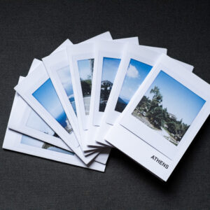 Photograph of 8 A7 zines fanned out on a dark grey fabric background. The top zine has the word 'ATHENS' in the bottom right corner and an Instax Square print on the cover that shows a temple in the Ancient Agora with plants below and blue sky above.