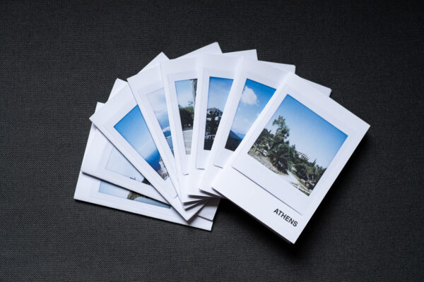 Photograph of 8 A7 zines fanned out on a dark grey fabric background. The top zine has the word 'ATHENS' in the bottom right corner and an Instax Square print on the cover that shows a temple in the Ancient Agora with plants below and blue sky above.