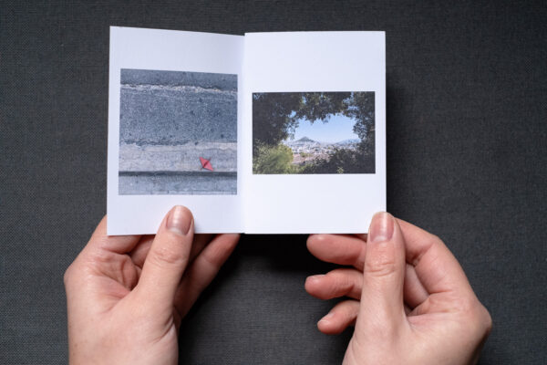 Photograph of an A7 zine on a dark grey fabric background being held by two hands. The zine is open to show 2 photographs, a red origami bird in the gutter and a hill shown through a gap in the foliage.