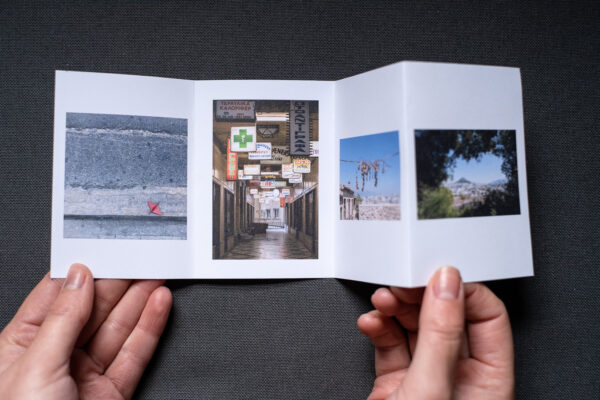 Photograph of an A7 zine on a dark grey fabric background, being held between two hands. The zine is unfolded and shows 4 photographs, a red origami bird in the gutter, lit signs in an alleyway, dried grapes hanging in front of blue sky and a hill shown through a gap in the foliage.