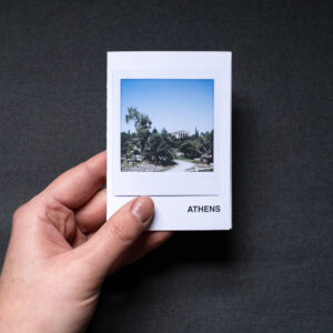 Photograph of an A7 zine held in a hand against a dark grey fabric background. The zine has the word 'ATHENS' in the bottom right corner and an Instax Square print on the cover that shows a temple in the Ancient Agora with plants below and blue sky above.