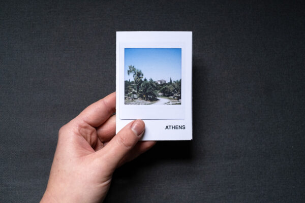 Photograph of an A7 zine held in a hand against a dark grey fabric background. The zine has the word 'ATHENS' in the bottom right corner and an Instax Square print on the cover that shows a temple in the Ancient Agora with plants below and blue sky above.