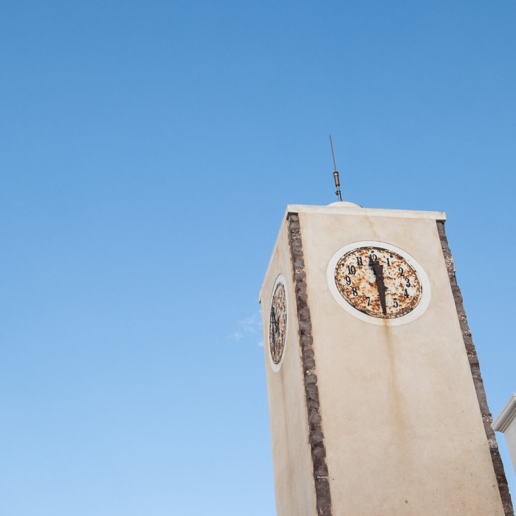 Photograph of a clock tower with a rusted clock and both hands pointing down, with rust having run down the tower. Blue sky behind.