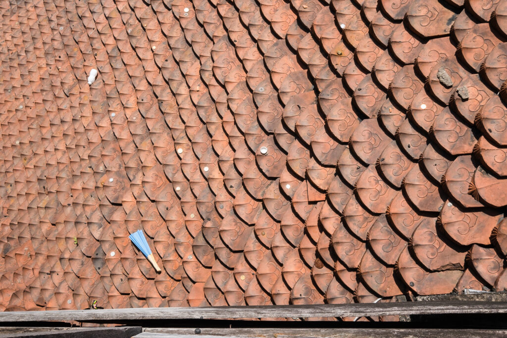 Photograph looking down on an ornate tiled roof with a blue paper folded fan that has fallen on to it to the left, a few coins and an empty Yakult bottle can also be seen.