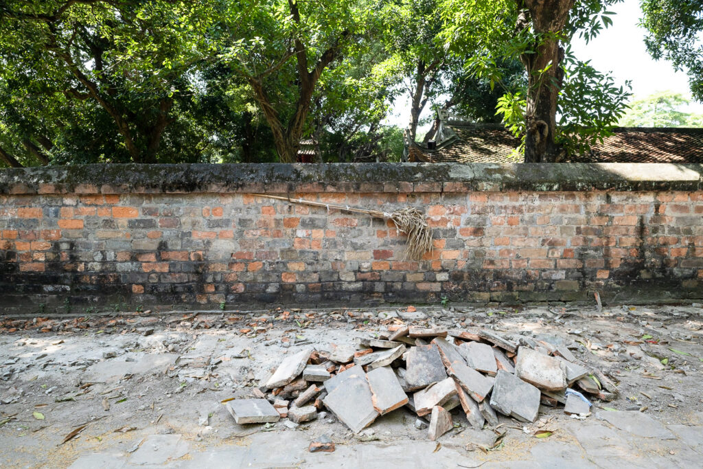 Photograph of a brick wall with a palm leaf broom hanging on it. Behind are trees and in front is a broken pavement with a pile of broken paving slabs.