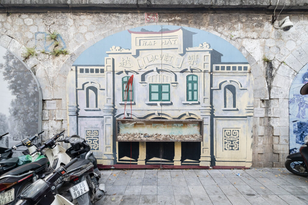 A photograph of a mural showing a painting of a building with 'Rap hat, lac viet' written on it. Lower down is a glass box that is half full of fallen leaves. Scooters can be seen to the left of the frame.