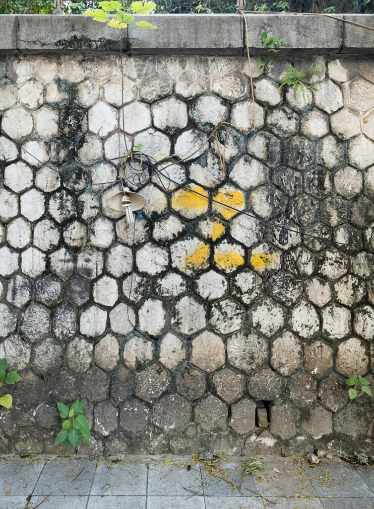 Photograph of a wall made with hexagonal bricks/tiles with a damaged lantern hanging on it, connected with lots of wires.