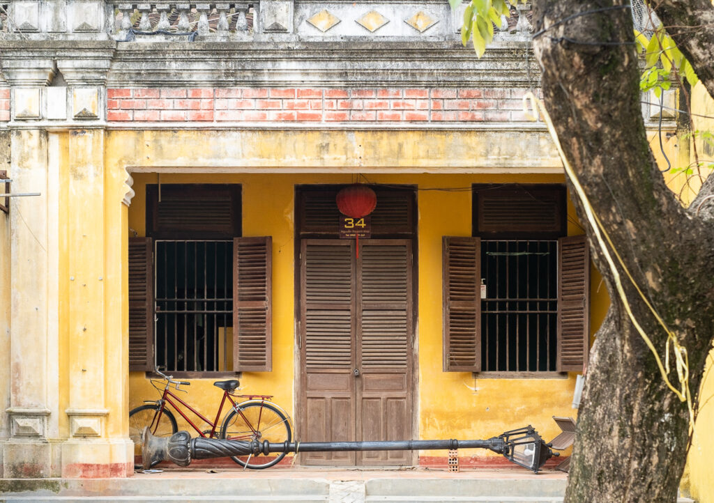 Photograph of a yellow building with shuttered doors and windows, a red bicycle and a large metal ornate lamp post lying down across the entrance way.