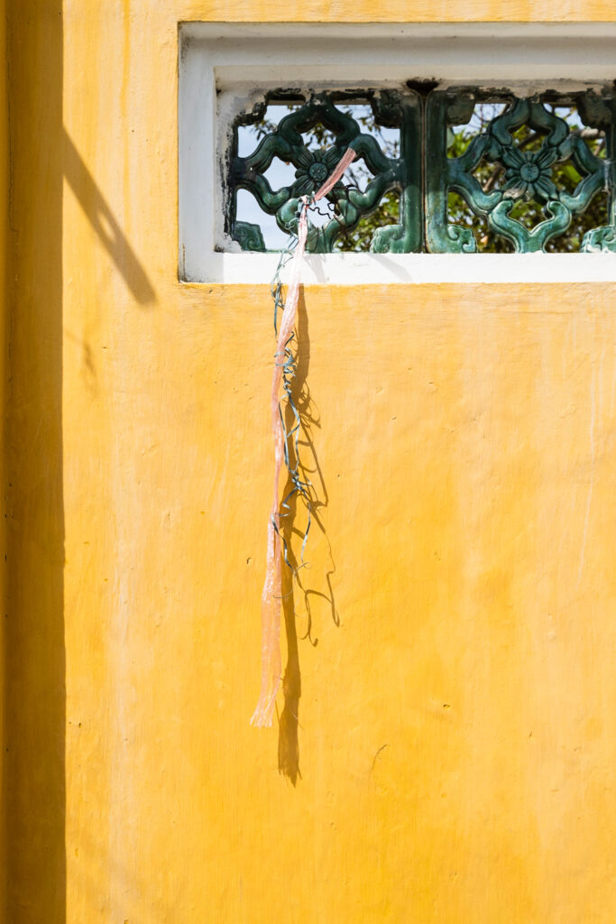 Photograph of a yellow wall with green decoration towards the top, with pink and green plastic ribbons tied to it and hanging down slightly tangled.