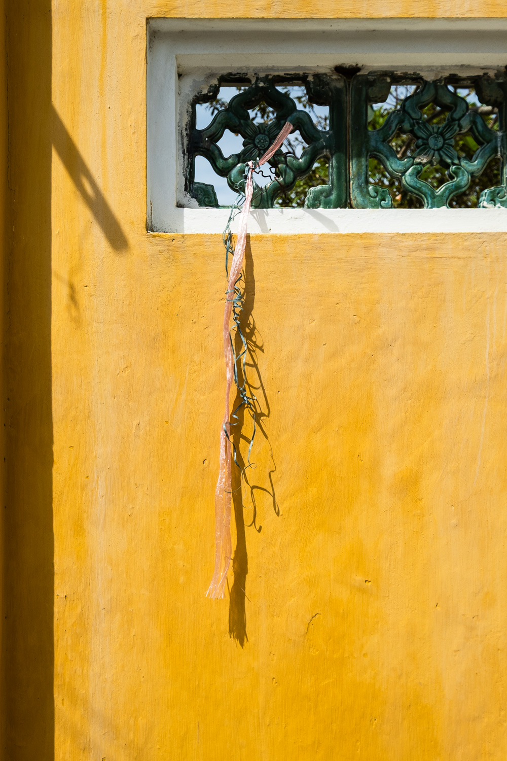 Photograph of a yellow wall with green decoration towards the top, with pink and green plastic ribbons tied to it and hanging down slightly tangled.