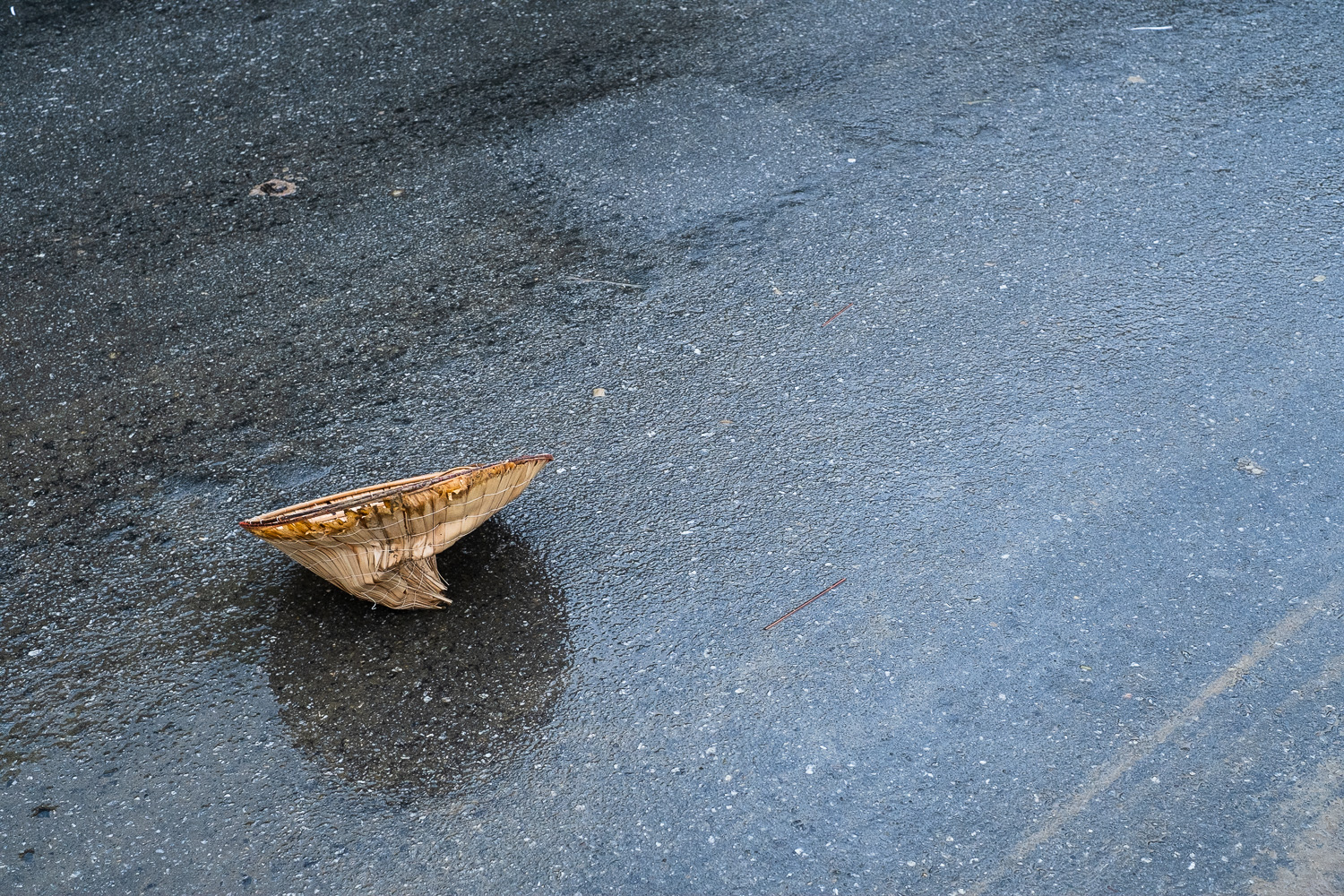 Photograph of a wet road with a nón lá (leaf hat) lying upside down and squashed towards the left of the frame.