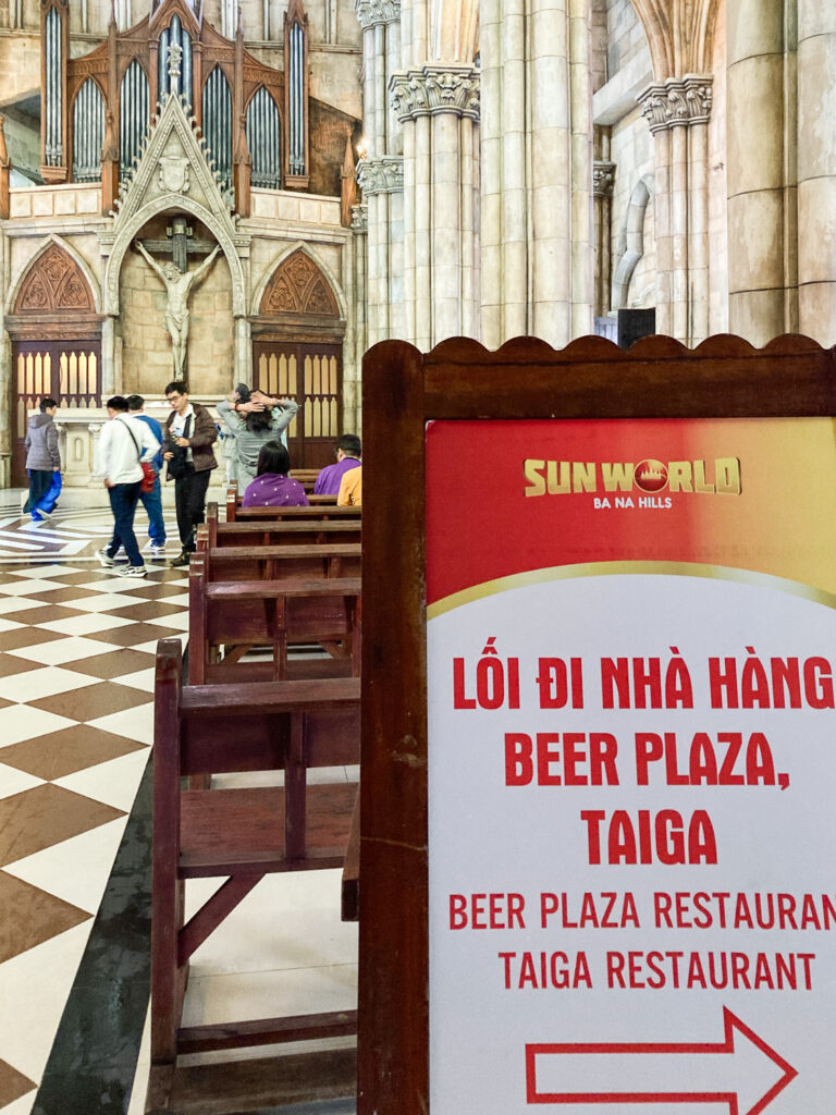 Interior of a replica Catholic church with organ and Jesus on the cross at the top left of the frame, with peole visible below, pews leading back to an advertising A-frame board in the foreground with 'Sun World, Ba Na Hills, (text in Vietnamese), Beer Plaza Restaurant, Taiga Restaurant' and an arrow pointing right.