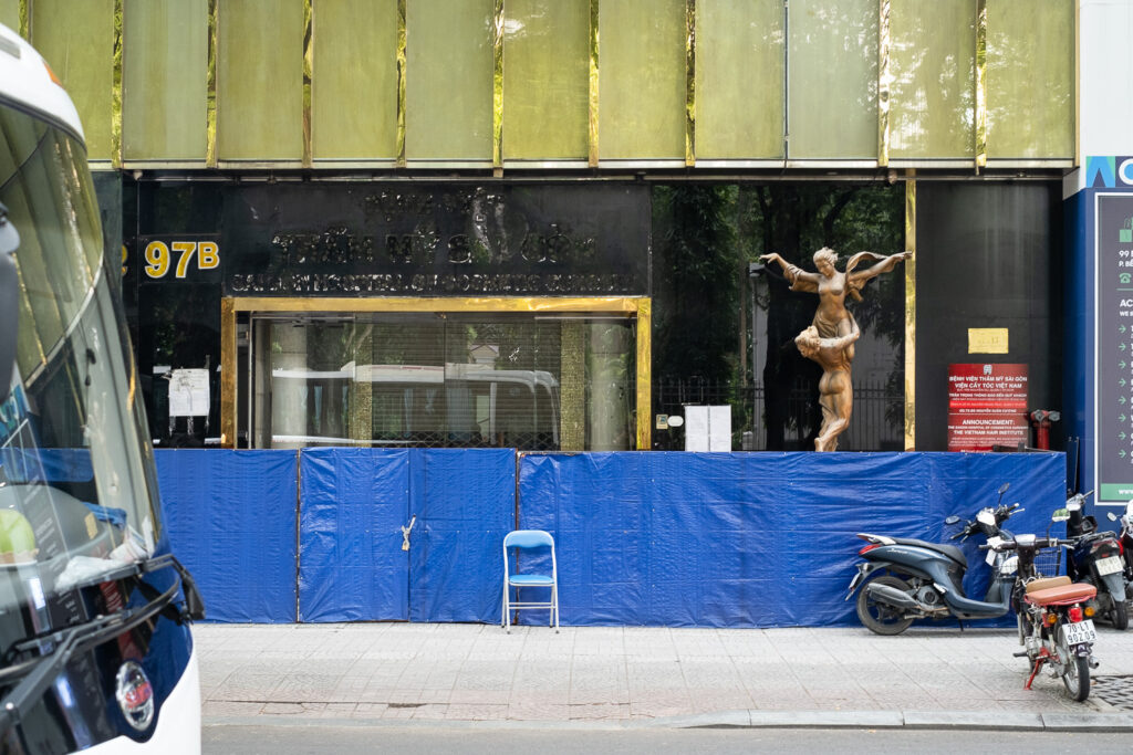 Photograph of a building with blue hoarding in front and a small blue chair, with a statue of a semi-naked couple to the right behind the hoarding, with one figure lifting the other, who has their arms wide as if flying. In the foreground there are scooters to the right on the pavement and the windscreen of a bus to the left.