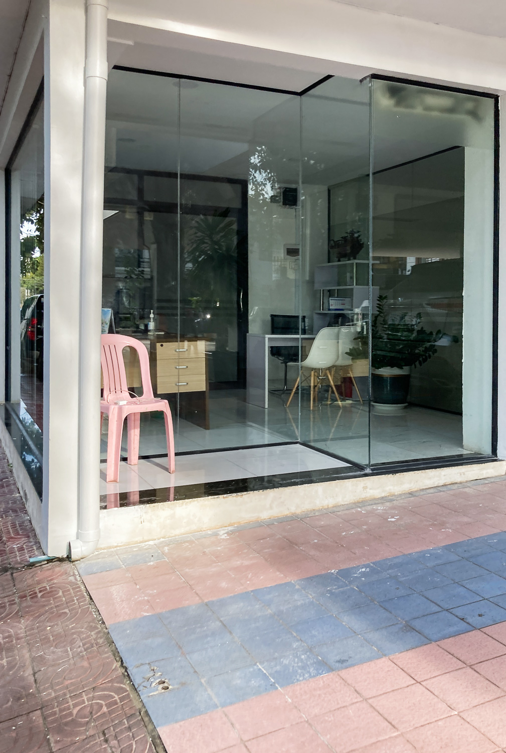 Photograph of a pink plastic garden chair in the entrance of a glass-fronted office, with pink and blue tiles on what appears to be a driveway in the foreground.