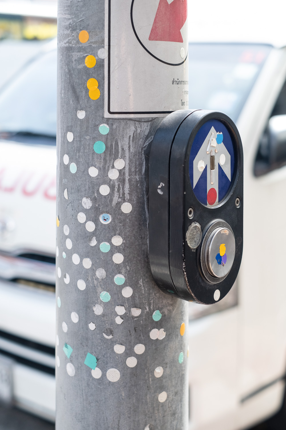 Photograph of a pole with circular stickers stuck on it (the stickers were proof of paying for entry to a location) and a box with buttons and an arrow pointing up, to press to cross the road). A white van is visible out of focus behind.