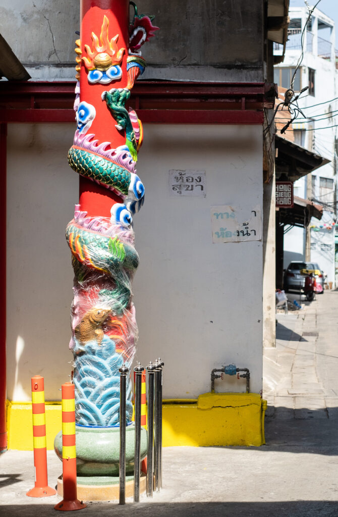 Photograph of an ornate pillar - red with a dragon snaking around it, a koi and waves at the bottom and cling film wrapped around the lower part. Bollards are around the bottom - red and yellow striped to the left and shiny metal to the centre. A wall with posters on is behind and a street can be seen to the right.