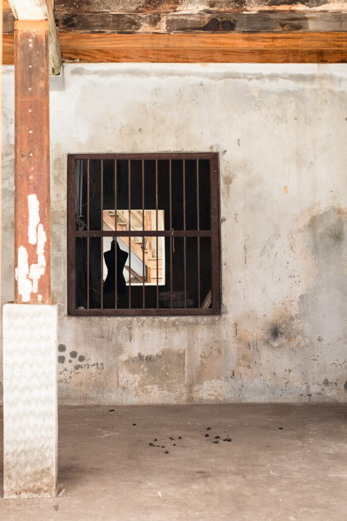 Photograph of a window with metal bars on it - behind it a dressmaker's dummy can be seen in silhouette with a staircase behind and in the foreground is a concrete wall and floor with some dirt on it and a wooden pillar with concrete base to the left.