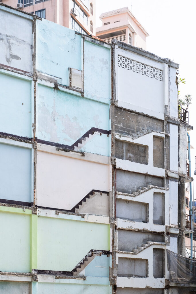 Photograh of the side of a building that has been partially knocked down, showing where stairs and doors were, and walls painted (top to bottom) light blue, slightly different shade of light blue, pink and green.