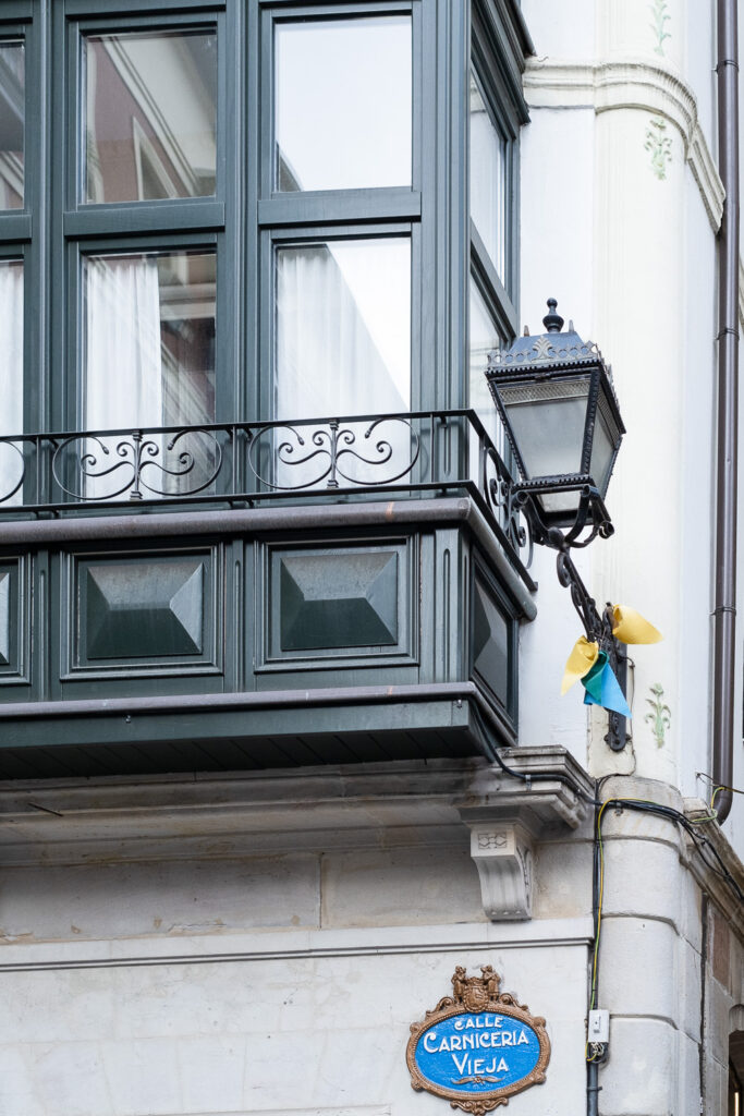 Photograph of a building with windows that look a bit balcony-like to the left, an ornate lantern to the right with yellow and blue bunting bunched up around it and an ornate sign saying 'Calle Carniceria Vieja' at the bottom of the frame.