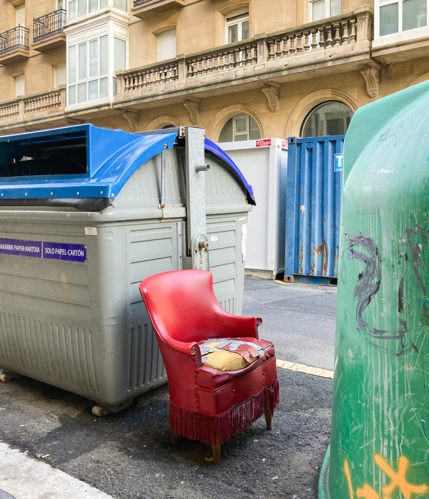 Photograph of a red armchair with  a seat that is ripped with the foam visible, it appears to have been fixed with tape many times in the past. The chair is on the roadside between bins and bottle recycling facilities, with ornate buildings visible on the opposite side of the road.