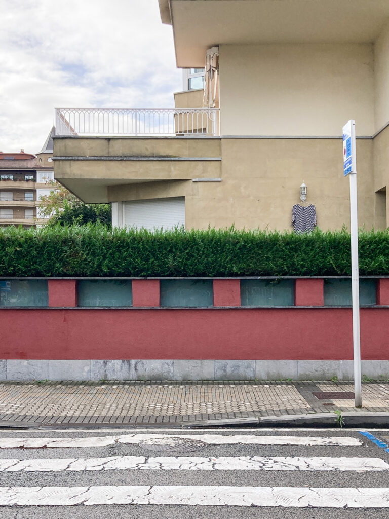 Photograph looking across a road with pedestrian crossing towards a red wall with neat hedge above and a cream coloured building behind. A tshirt is hanging from a coathanger attached to a lantern on the wall of the bulding, which looks a little like a person.