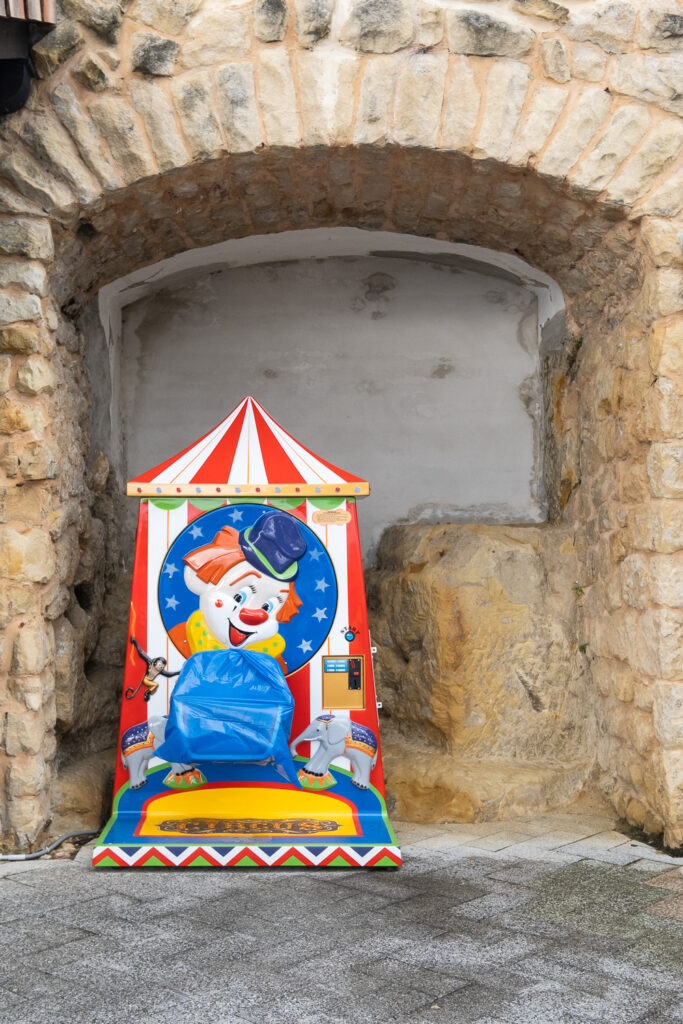 Photograph of a child's ride with circus and clown decoration with a seat covered by a blue plastic sheet. The ride is positioned by an alcove in an old wall.