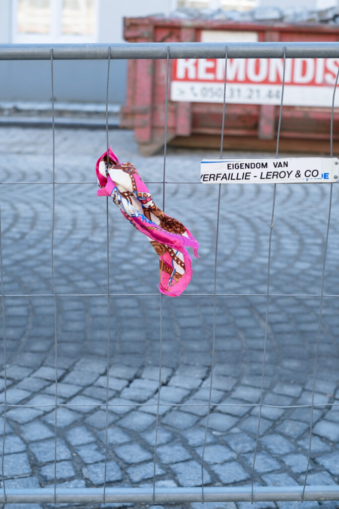 Photograph of a pink, brown and cream scarf tied to a thin metal temporary fence and blowing in the windw. Behind is a cobbled pavement and a skip out of focus in the background.