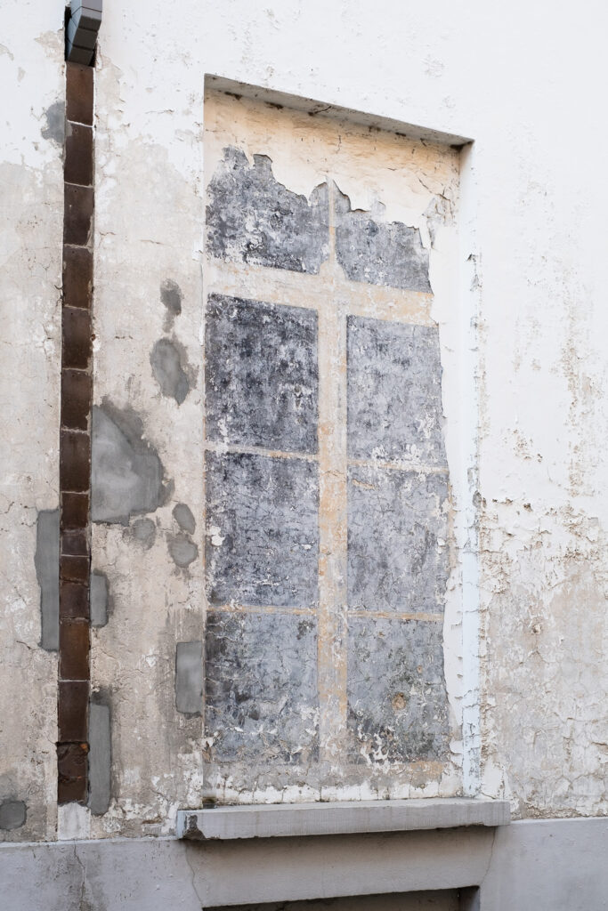 Photograph of a wall with what look looks like a filled in window that has been painted to look like a window and now the plaster and paint is flaking off. There is a line of bricks or tiles to the left.