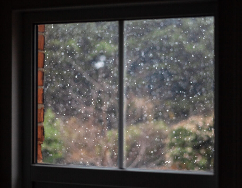 Photograph of snowflakes falling outside a window, with out of focus garden behind and the edge of a brick wall to the left, the dark interior around the edges.