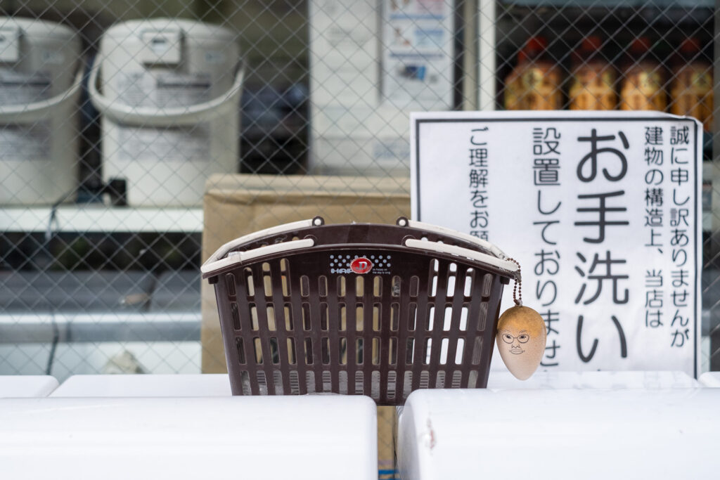 Photograph of a brown plastic shopping basket on a white surface with a keyring in the shape of a lifesize egg with a face of a person with glasses on it. Behind is a sign in Japanese and shelves with products on behind a reinforced window.