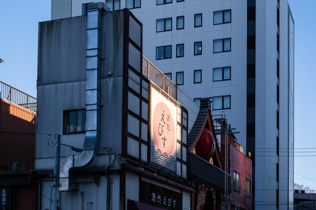 Photograph of buildings in Tokyo as the sun sets. The sun is shining on a sign with an image of a sun and waves and Japanese writing on it. The buildings around this are darker and blue with the fading light.