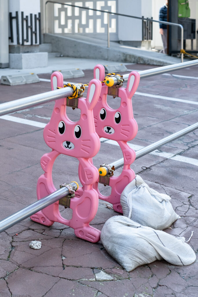 Photograph of barriers with pink plastic rabbit characters at the ends of them (centre of the image). There are sandbags to their right, the ground beneath is cracked beneath and parking spaces and buildings are visible behind.