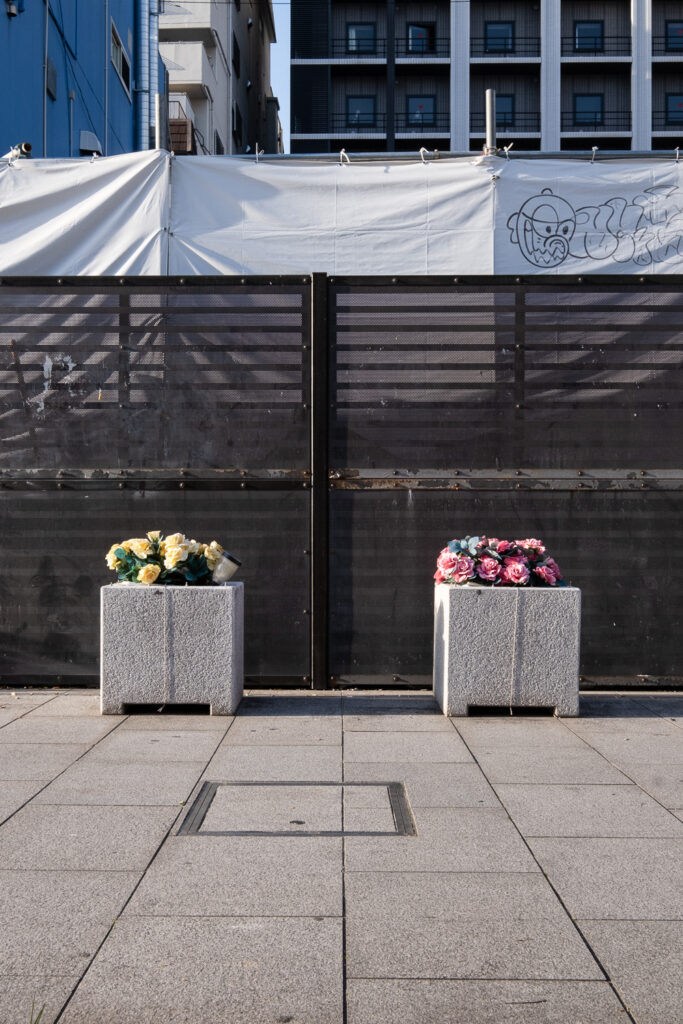 Photograph of two concrete planters filled with fake flowers - yellow on the left and red on the right. A paved pavement is in the foreground and metal fencing and hoarding with some grafitti is visible behind and buildings even further behind.