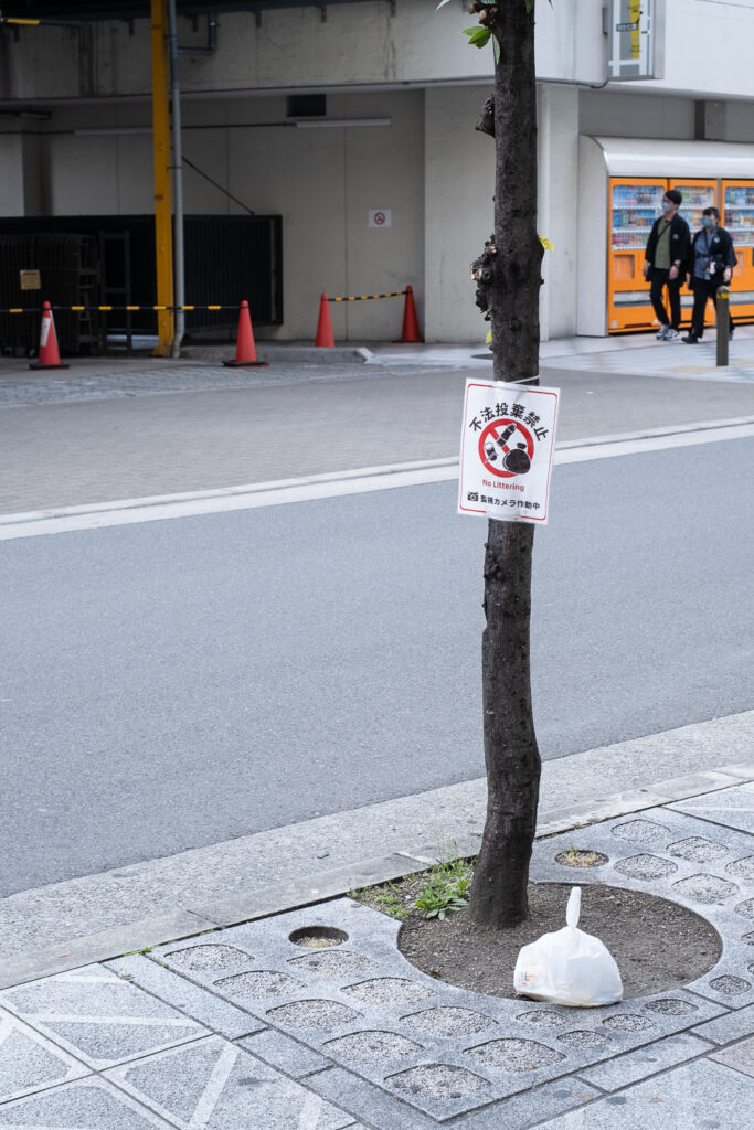 Photograph of a tree with a 'no littering' sign on it, with a bag of rubbish left underneath it, which looks almost exactly like one of the symbols in the 'no littering' sign (which also shows a bottle and takeaway cup crossed out). The tree is in a cutout area of pavement and a road, building, cones and two people are visible behind.