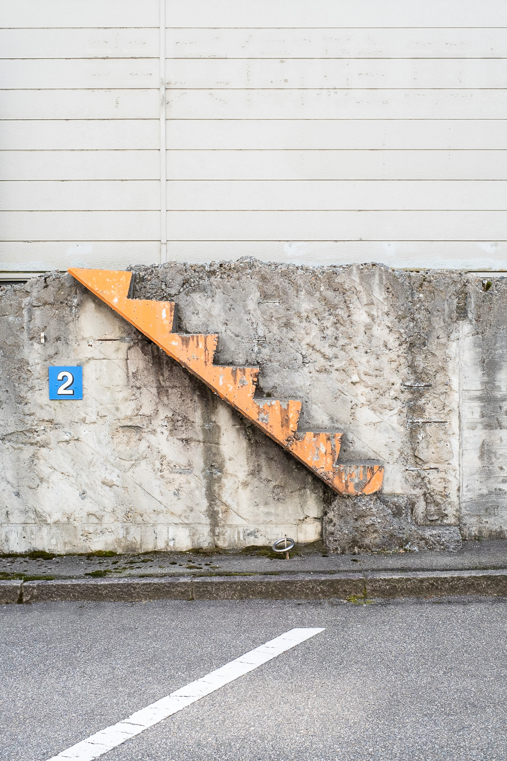 Photograph of a concrete wall with the profile of steps that has been painted orange going upwards from the pavement to the top of the rough wall right to left, a smooth wall with horizontal lines along it above and behind, a blue sign with '2' on it to the left and a diagonal white painted line on the road in front indicating parking spaces.