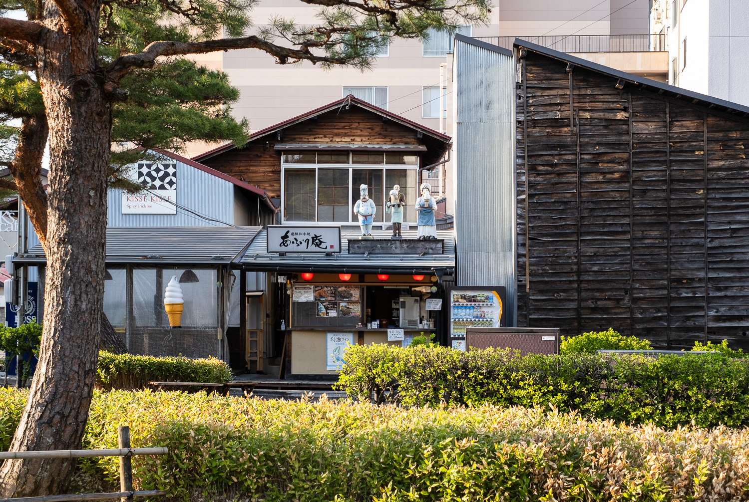 Restaurant with plastic models of ice cream in a cone, a chef and waiting staff to advertise it. Takayama, Gifu Prefecture, Japan. Ice cream, beer, ice cream. Photograph looking over hedges and a tree towards buildings with a large plastic ice cream cone to the left, a shop front with 3 statues of people in chef/serving outfits holding a large glass of beer and an ice cream cone on a tray in the centre and the side of a wooden building to the right.