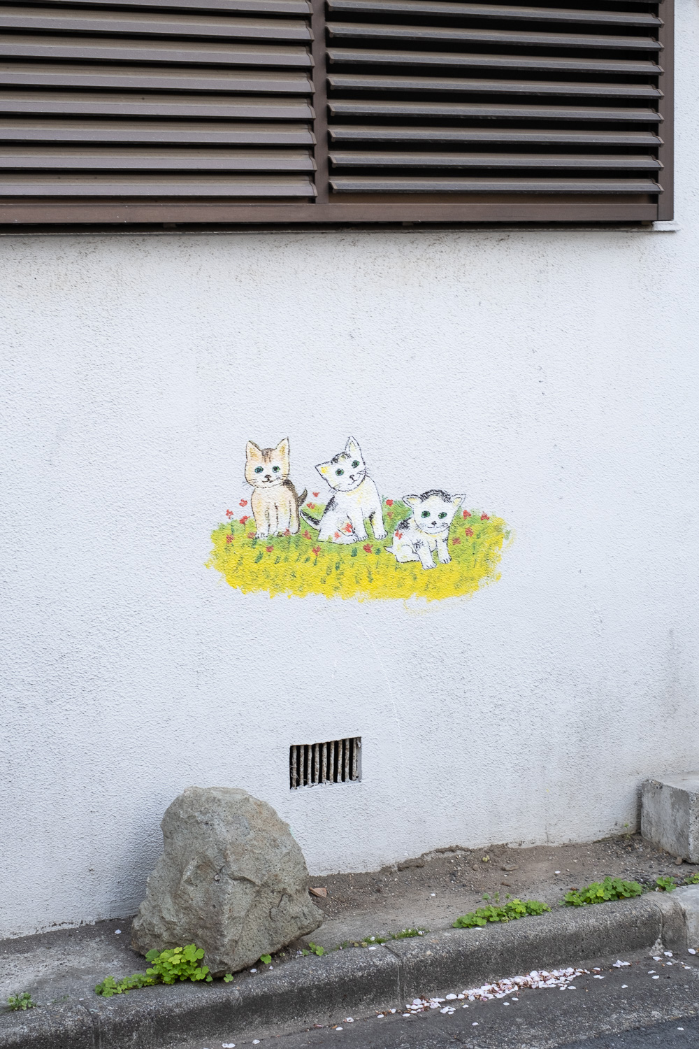 Photograph of a white wall with brown shutters towards the top and pavement with a large rock, weeds and fallen blossom below. In the middle of the wall is a painted mural of 3 kittens sitting on grass with red flowers around them.