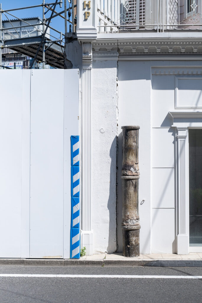 Photograph across a road of hoarding to the left with a section of blue and white striped plastic and scaffolding visible behind, part of a white painted building to the right with a pillar made to look like over-sized bamboo, with a balcony just visible above and part of doorway to the right.