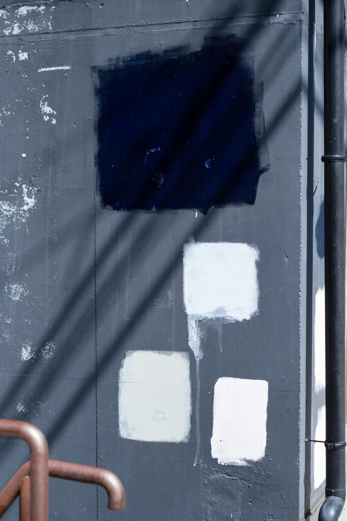 Photograph of a grey wall with squares painted in black, very light blue, very light green and very light pink. A drainpipe can be seen to the right and shadows of wires go diagonally from the bottom left to top right corners.