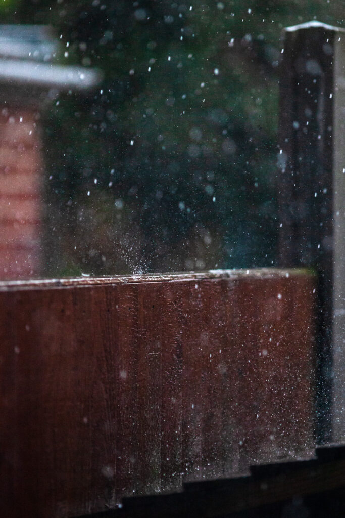 Photograph of heavy rain taken at a fast shutter speed and selective focus to show droplets bouncing of a brown wooden fence. Trees, a building and a concrete fence post can be seen out of focus in the background.