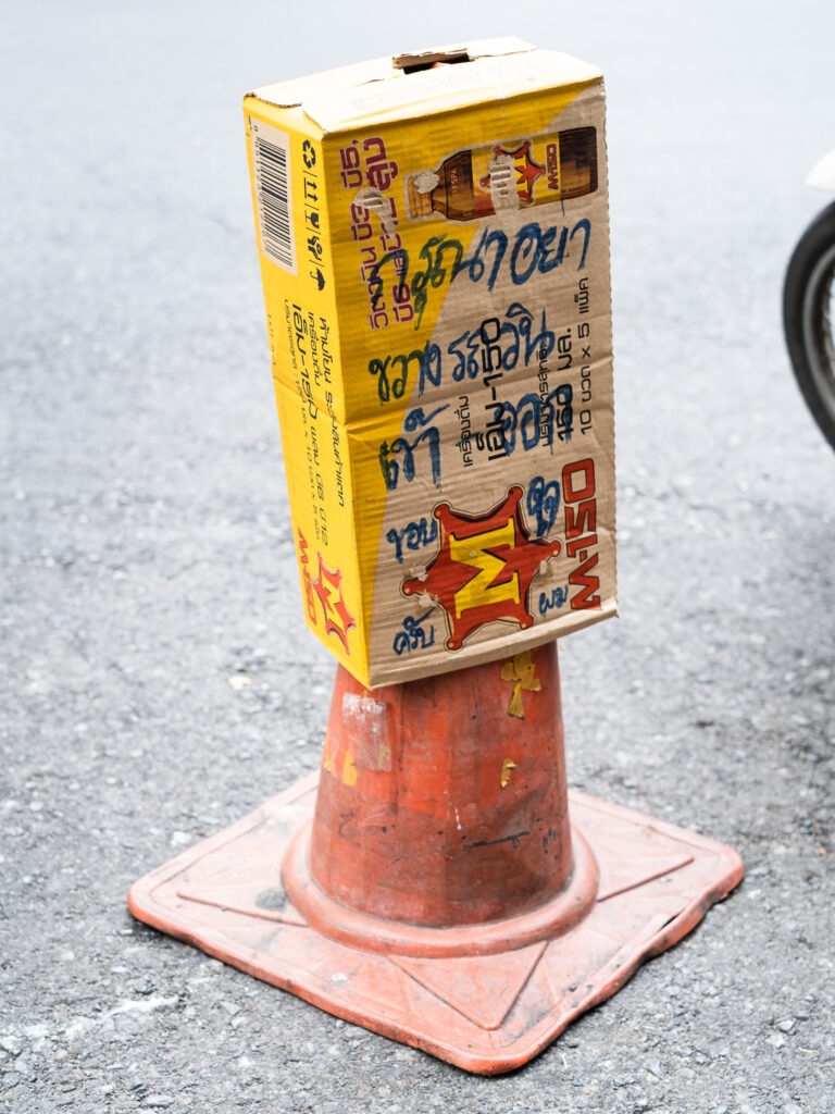 Photograph of an orange plastic traffic cone on a road with a box with 'M-150' and an image of a bottle on it, with handwriting in Thai that says 'Please don’t park your cars in front of the entrance and exit ways of the motorbike pick-up point. Thank you.'